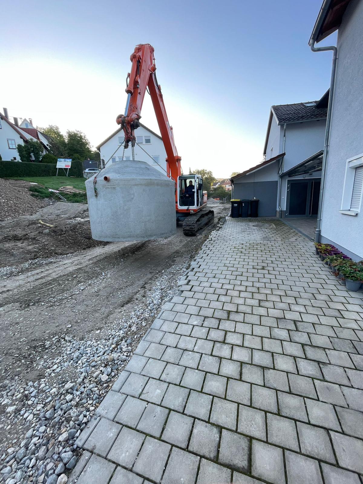 Bagger hebt eine Betonzisterne neben der fertigen Pflasterzufahrt eines Wohnhauses ein